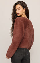 Woman looking over her shoulder while wearing a brown fuzzy sweater and black jeans against a neutral background