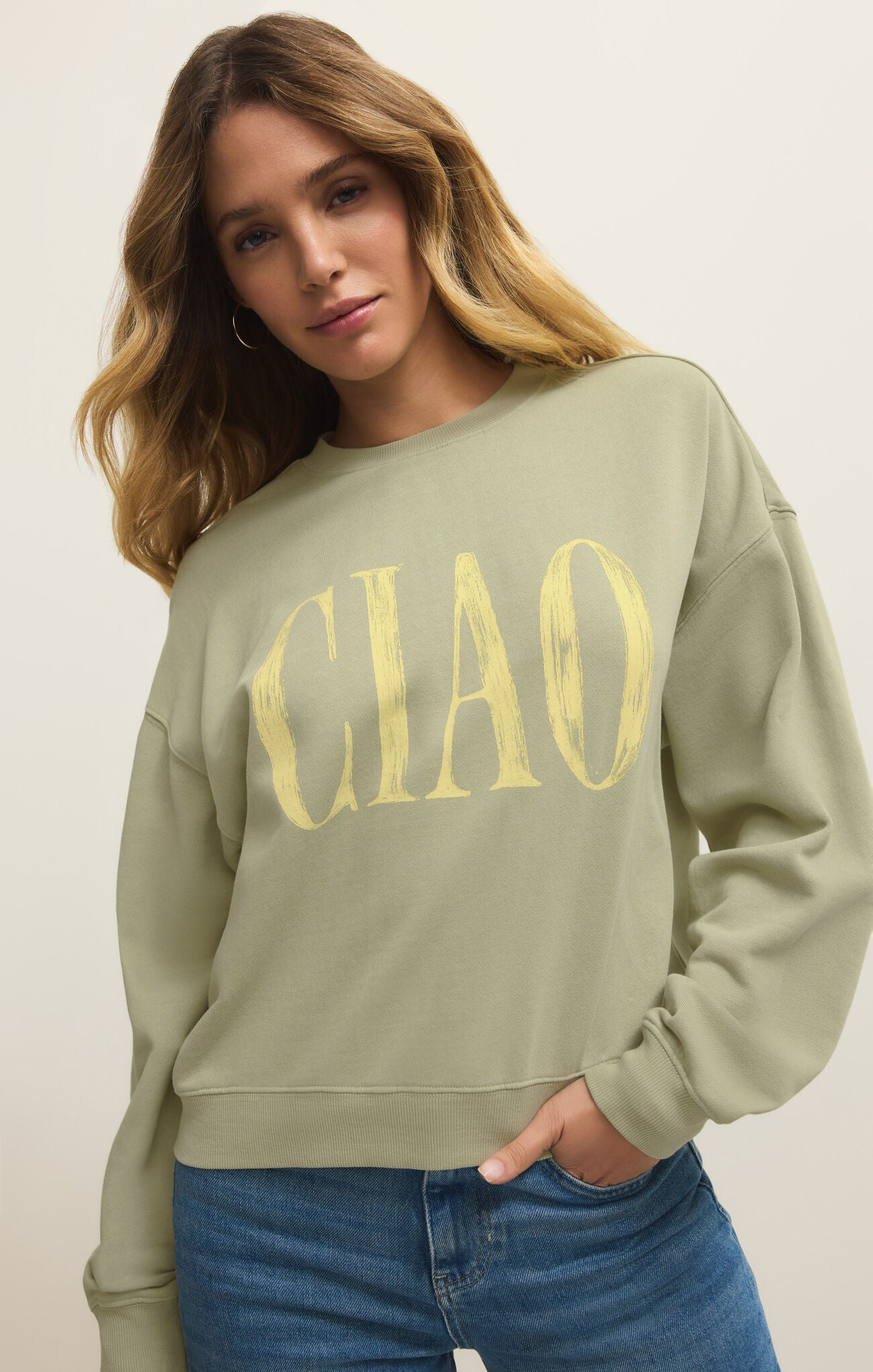 Woman with wavy blonde hair wearing a light green sweatshirt with yellow "CIAO" text and blue jeans standing against a neutral background