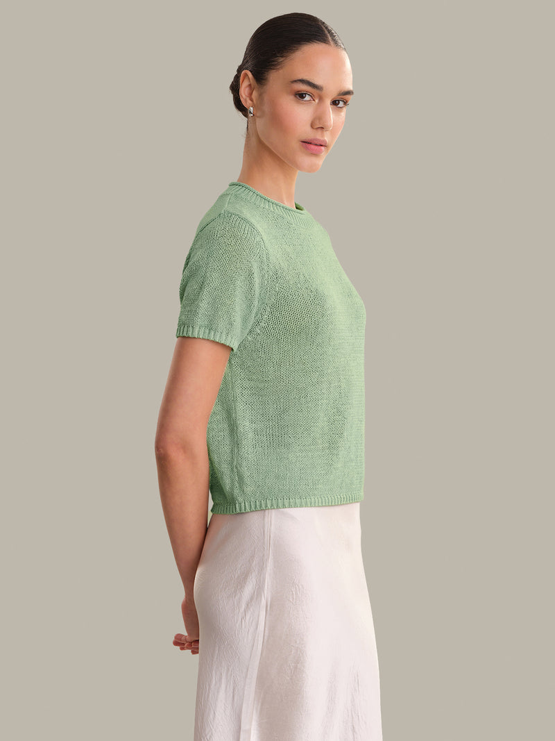 Model wearing a silt green Faye Rolled Edge Sweater Tee by 525 america paired with a light skirt against neutral backdrop