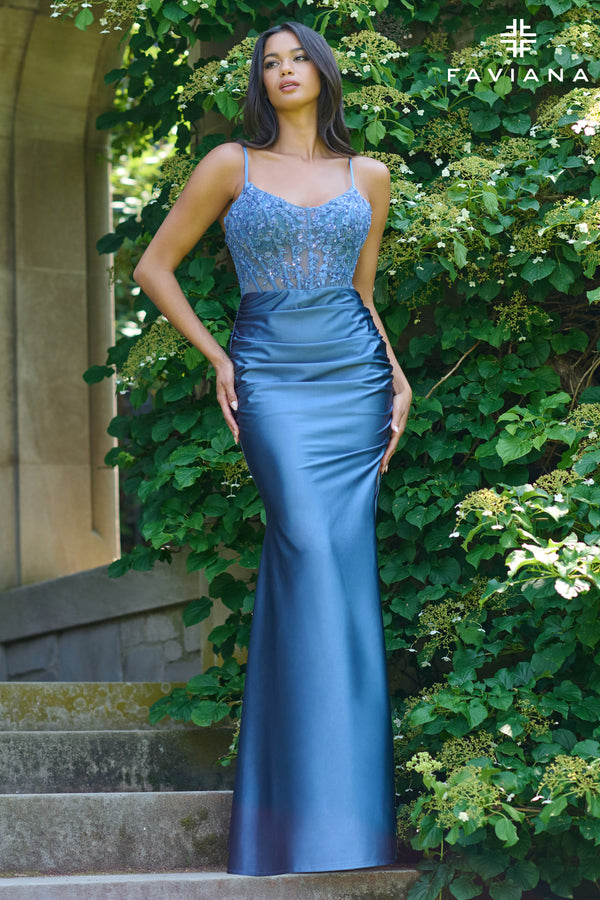 Woman with long dark hair wearing a steel blue fitted prom dress with lace bodice standing outdoors by greenery