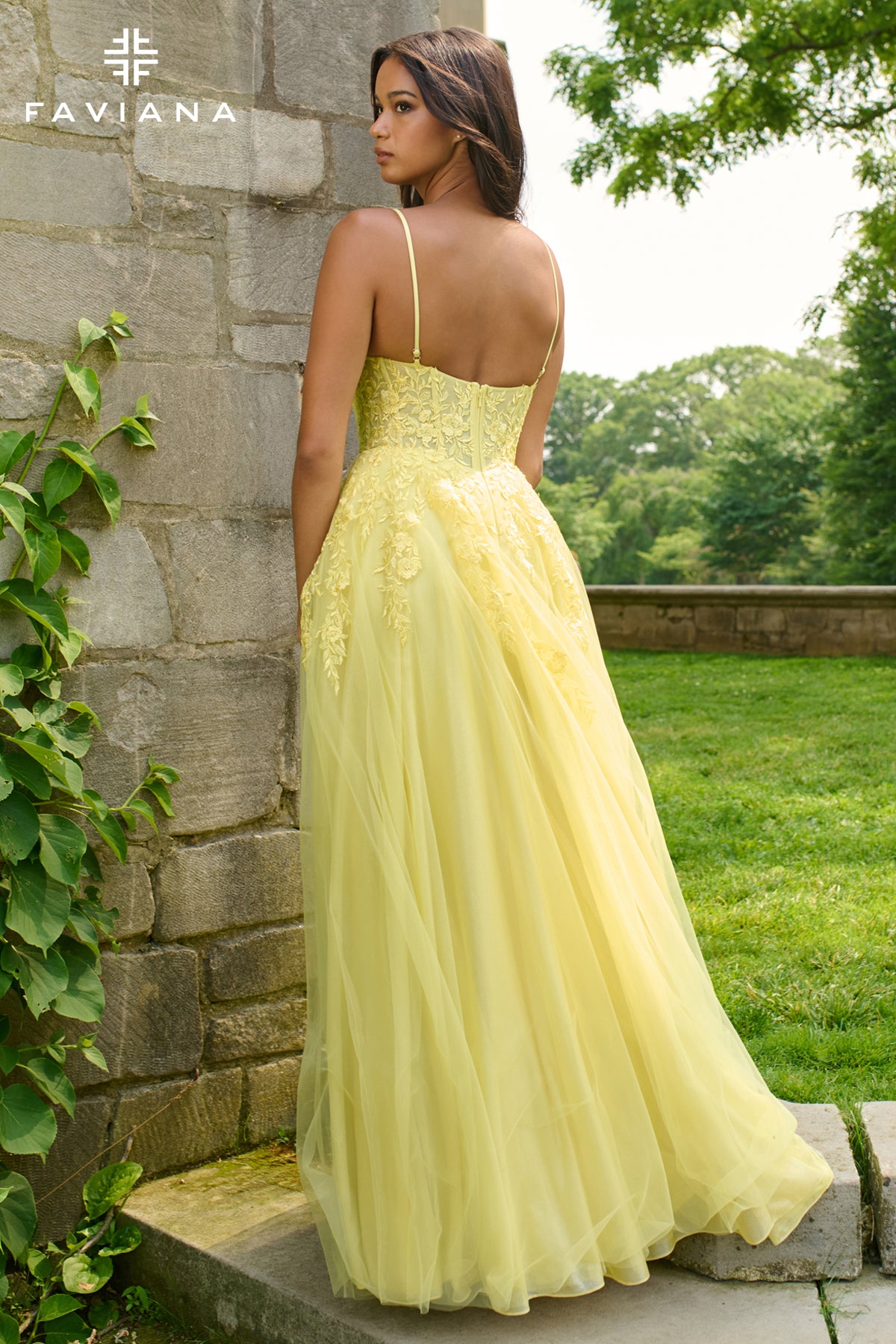 Woman in a yellow evening gown standing against a stone wall with greenery in the background, FAVIANA brand visible.
