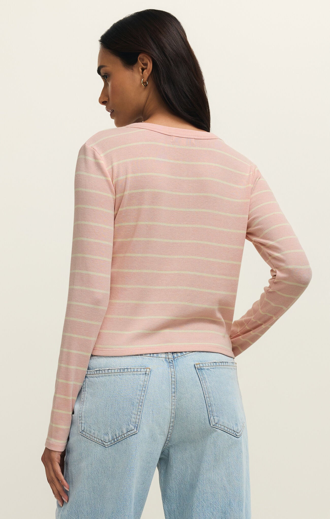 Woman with dark hair wearing a washed pink and cream striped long-sleeve Henley top and light blue jeans facing away