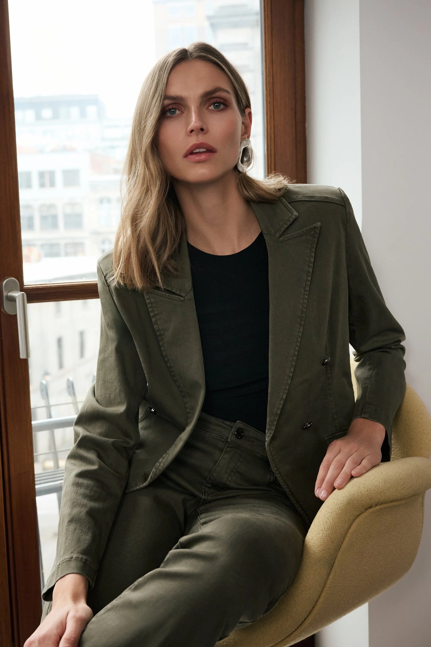JOSEPH Ribkoff Avocado Blazer in olive green worn with a dark top and matching trousers while seated in an armchair