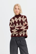 Blonde woman wearing a burgundy and beige argyle sweater with a high neck and black denim jeans standing with arms crossed