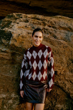 Woman wearing a burgundy and beige argyle sweater and black leather skirt standing against a large rock background