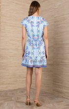 Back view of a woman wearing a floral Hale Bob Blue Dulce Dress in a light blue shade with ruffle hem and gold heels.