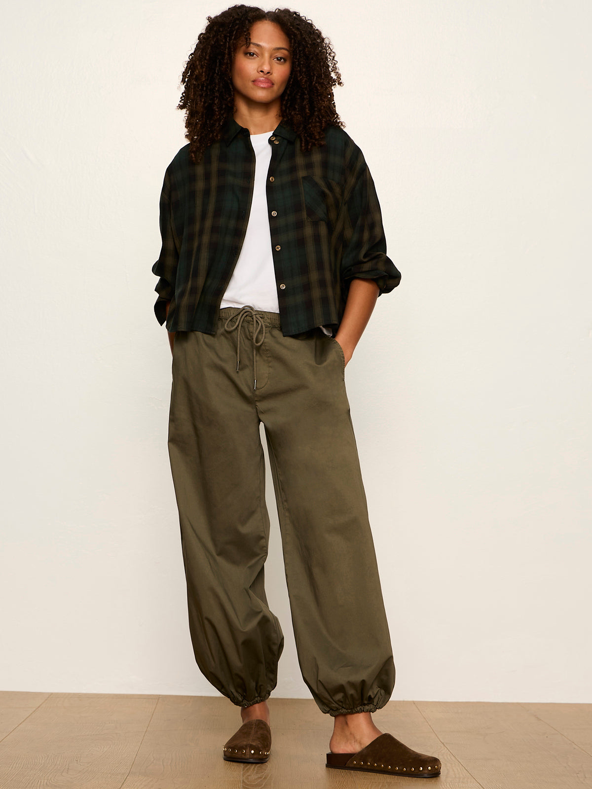 Woman with curly hair wearing a dark plaid shirt over a white top, olive drawstring pants, and brown slip-on shoes