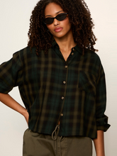 Woman with curly hair wearing black sunglasses and a dark green plaid button-up shirt with rolled sleeves