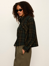 Woman wearing dark green and black plaid shirt with olive pants and black sunglasses against a plain background