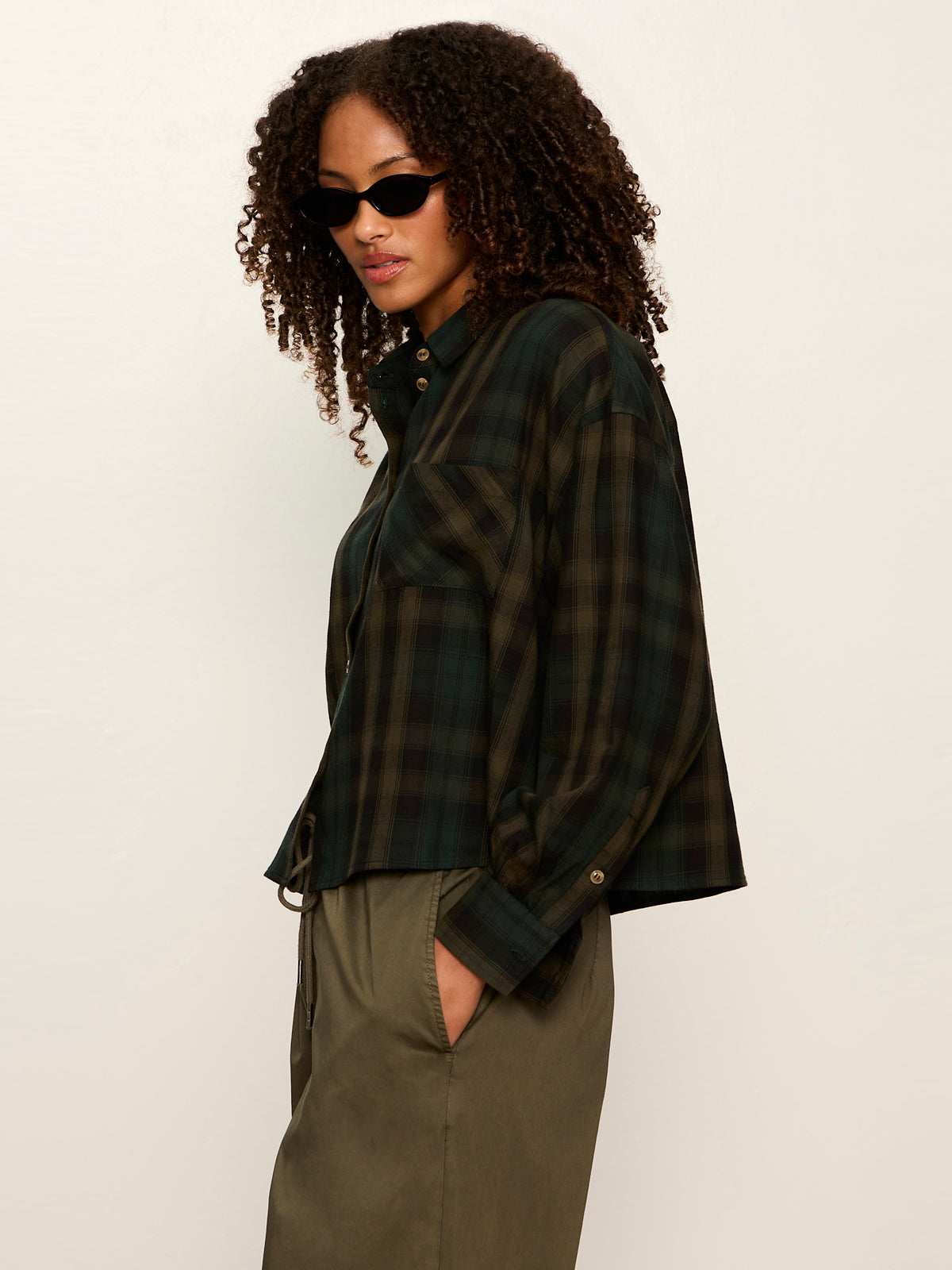 Woman wearing dark green and black plaid shirt with olive pants and black sunglasses against a plain background