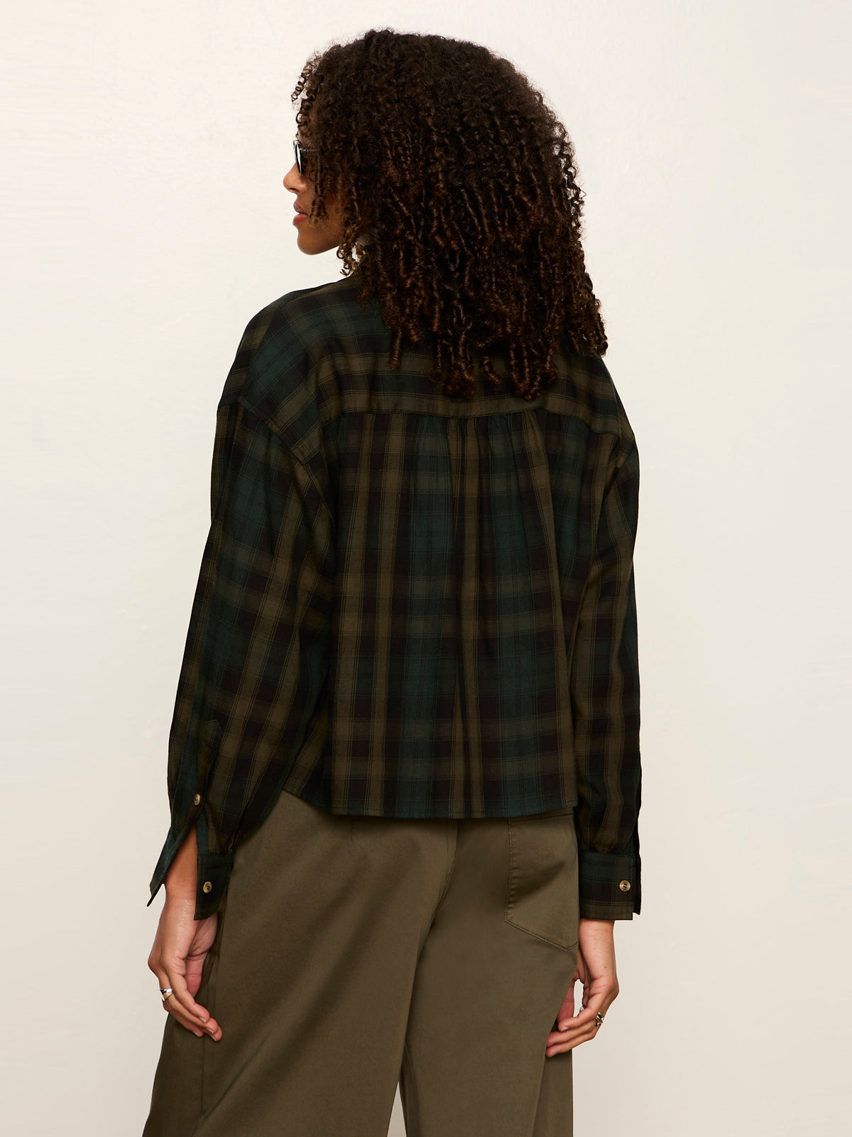 Woman with curly hair wearing a dark green and black plaid long-sleeve shirt and olive green pants facing away from camera.