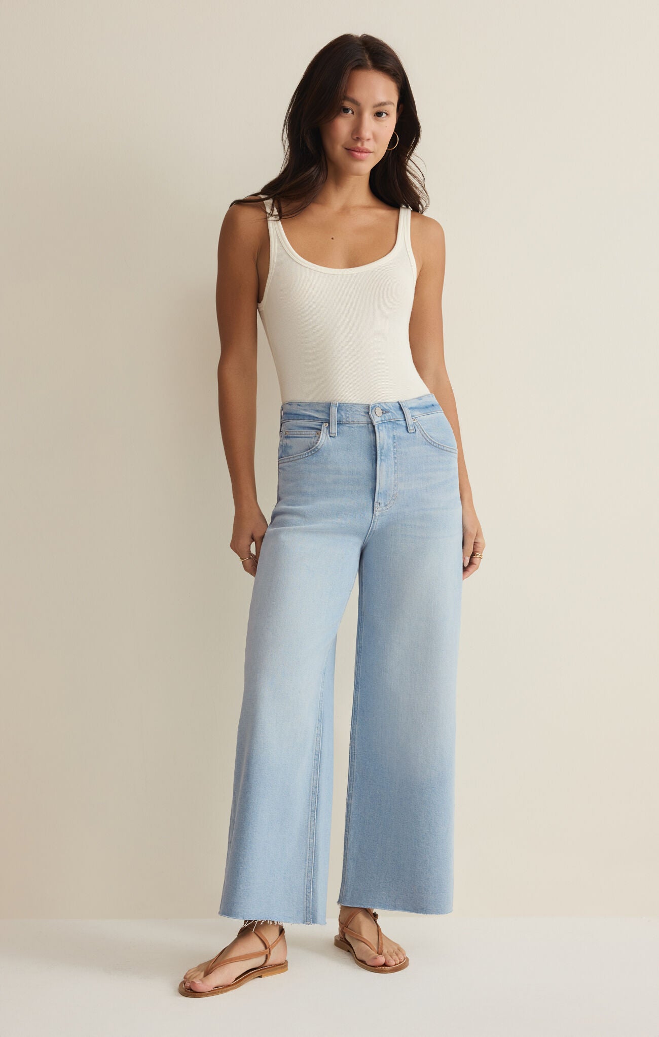 Woman with long dark hair wearing a white tank top and light blue high-rise wide-leg cropped jeans standing against a plain background