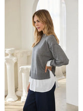 Woman wearing a gray sweater over a white shirt and navy trousers in a bright classical interior