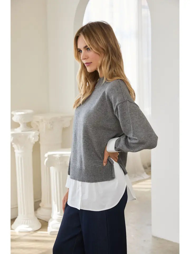 Woman wearing a gray sweater over a white shirt and navy trousers in a bright classical interior