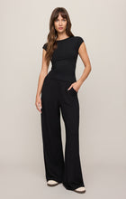 Woman wearing a Black Soft Trace Second Skin Top by z supply with relaxed black pants and white shoes standing against beige background