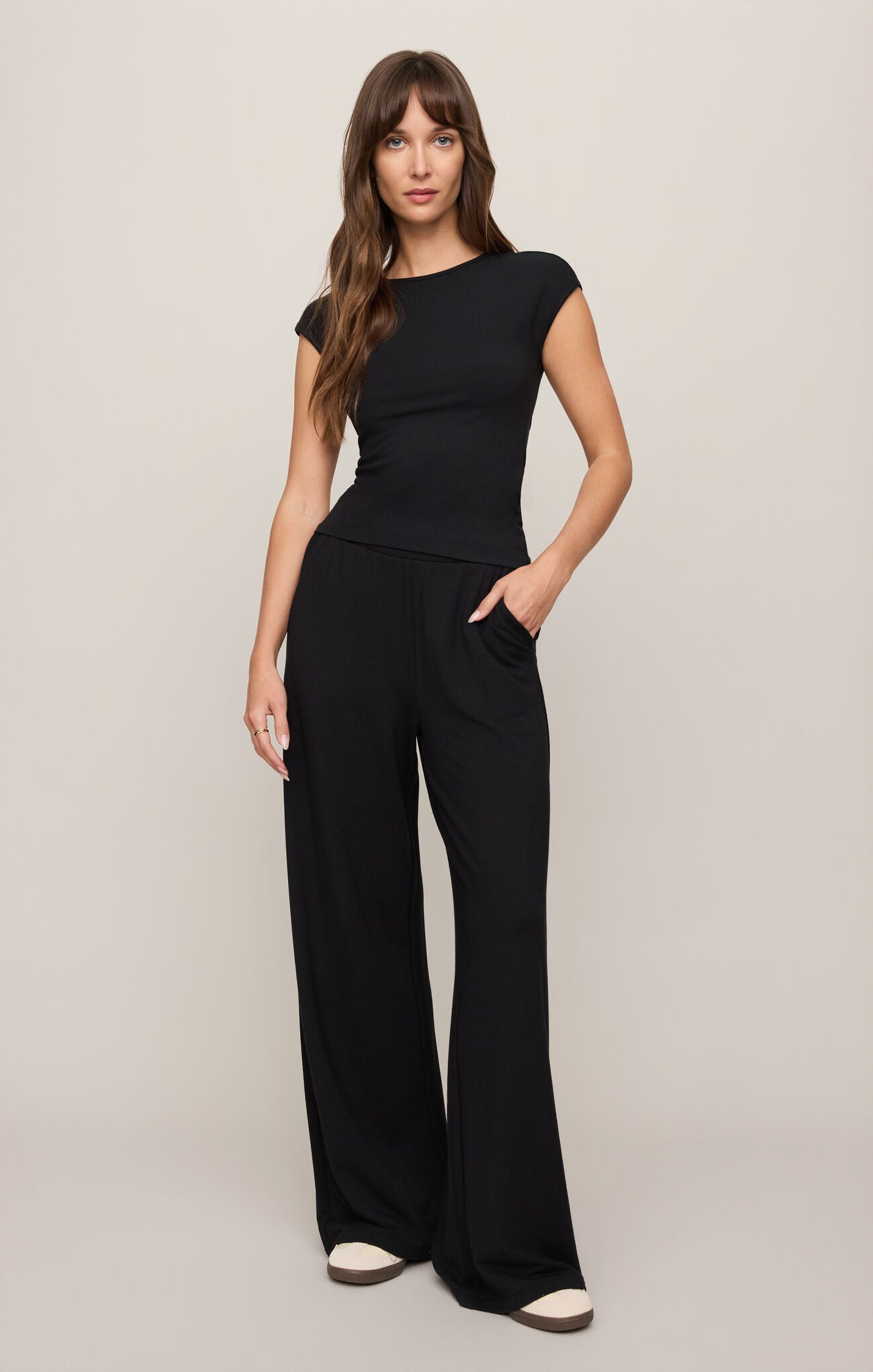Woman wearing a Black Soft Trace Second Skin Top by z supply with relaxed black pants and white shoes standing against beige background