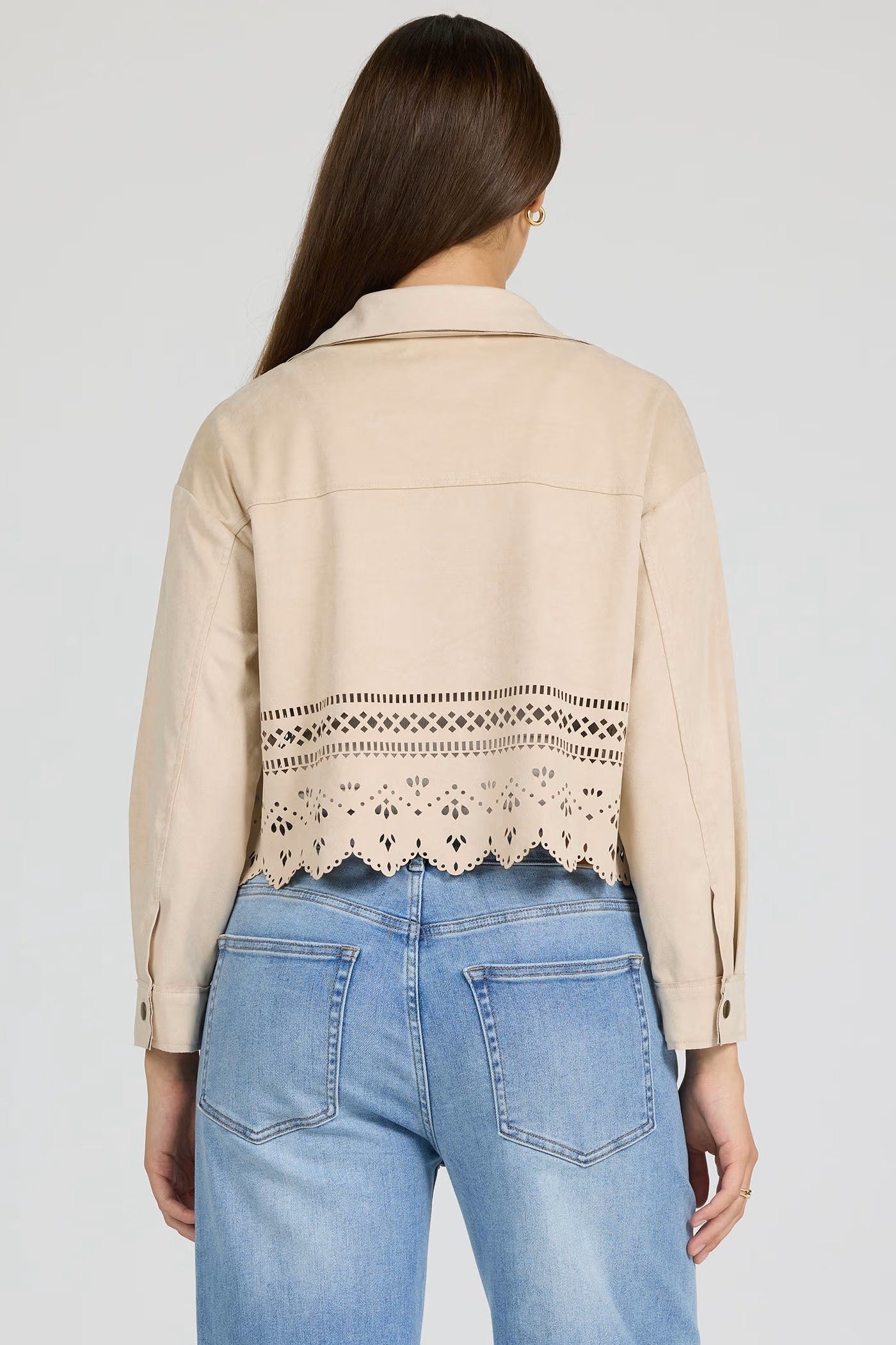Back view of a woman wearing a beige Dear John Retta Jacket with laser-cut detailing above the hem and light blue jeans