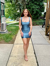 A woman wearing an aqua blue mini dress and nude high heels walking on a concrete sidewalk outdoors.