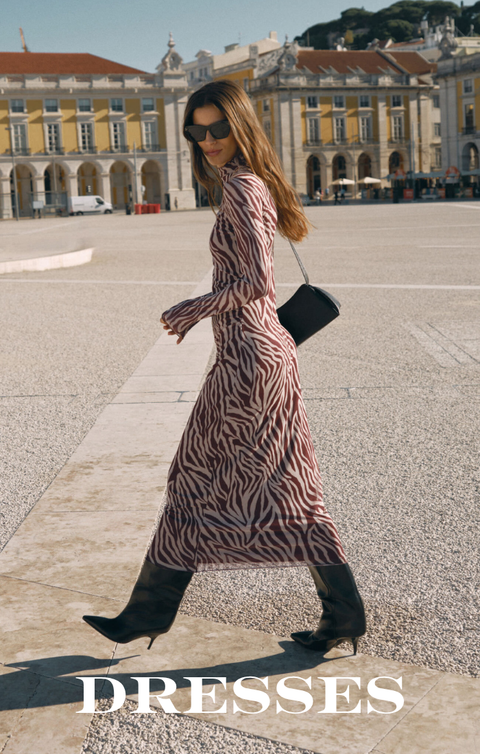 A woman walking in a large sunlit plaza wearing a long-sleeve midi dress with a pink and beige zebra-like pattern, black pointed boots, sunglasses, and carrying a small black handbag. Historic buildings are visible in the background, and the word 'DRESSES' appears at the bottom.