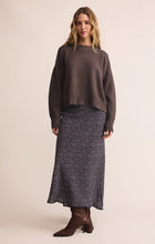 Woman with wavy blonde hair wearing a dark brown sweater and black floral maxi skirt with brown ankle boots standing against a plain background