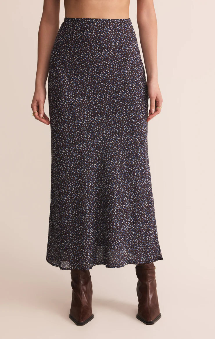 Woman wearing a dark floral maxi skirt with a fitted waist and flowy hem paired with brown boots standing against a beige backdrop