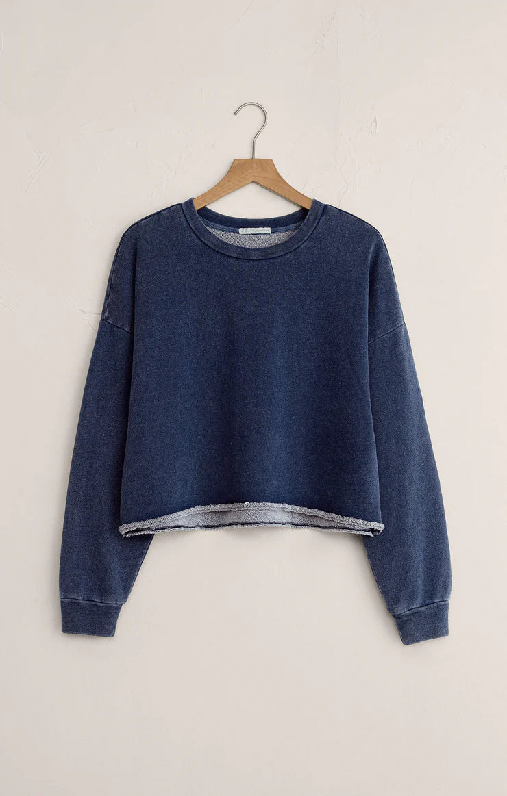 Blue knit denim sweatshirt with raw edge hem and long sleeves hanging on a wooden hanger against a beige wall