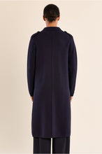Back view of a person wearing a long dark navy coat with shoulder details and matching trousers