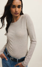 Woman with long dark hair wearing a natural heather long-sleeve crewneck shirt and blue jeans against a plain background