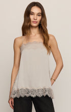 Woman wearing a beige lace trimmed camisole and black pants against a neutral background