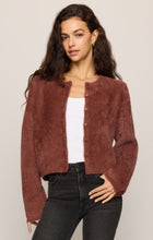 Woman wearing a brown cardigan over a white top with black jeans against a neutral background