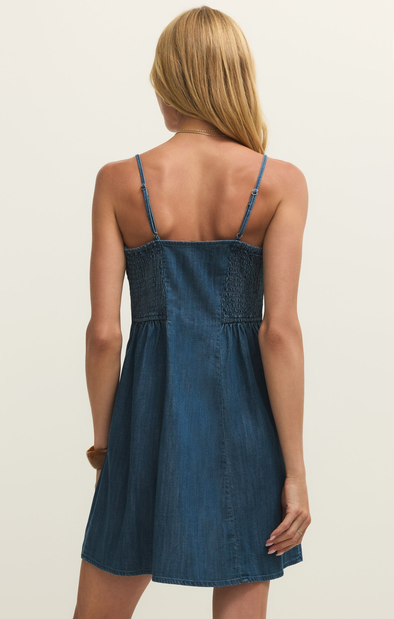 Back view of a woman wearing the Medium Indigo Angel Eyes Denim Mini Dress by z supply with adjustable straps and smocked bodice