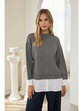 Woman wearing a gray sweater layered over a white shirt and dark trousers indoors