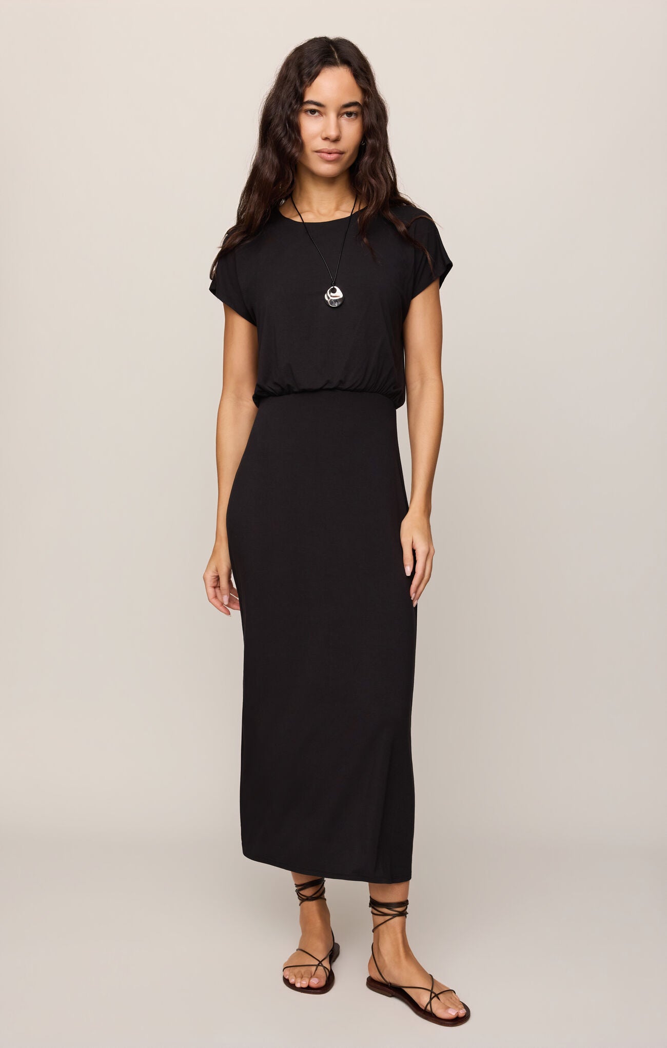 Woman wearing a Black Anika Midi Dress by z supply with short sleeves and gathered waist, styled with sandals and necklace