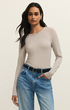 Woman with dark hair wearing a natural heather long-sleeve crew neck top and blue jeans standing against a neutral background