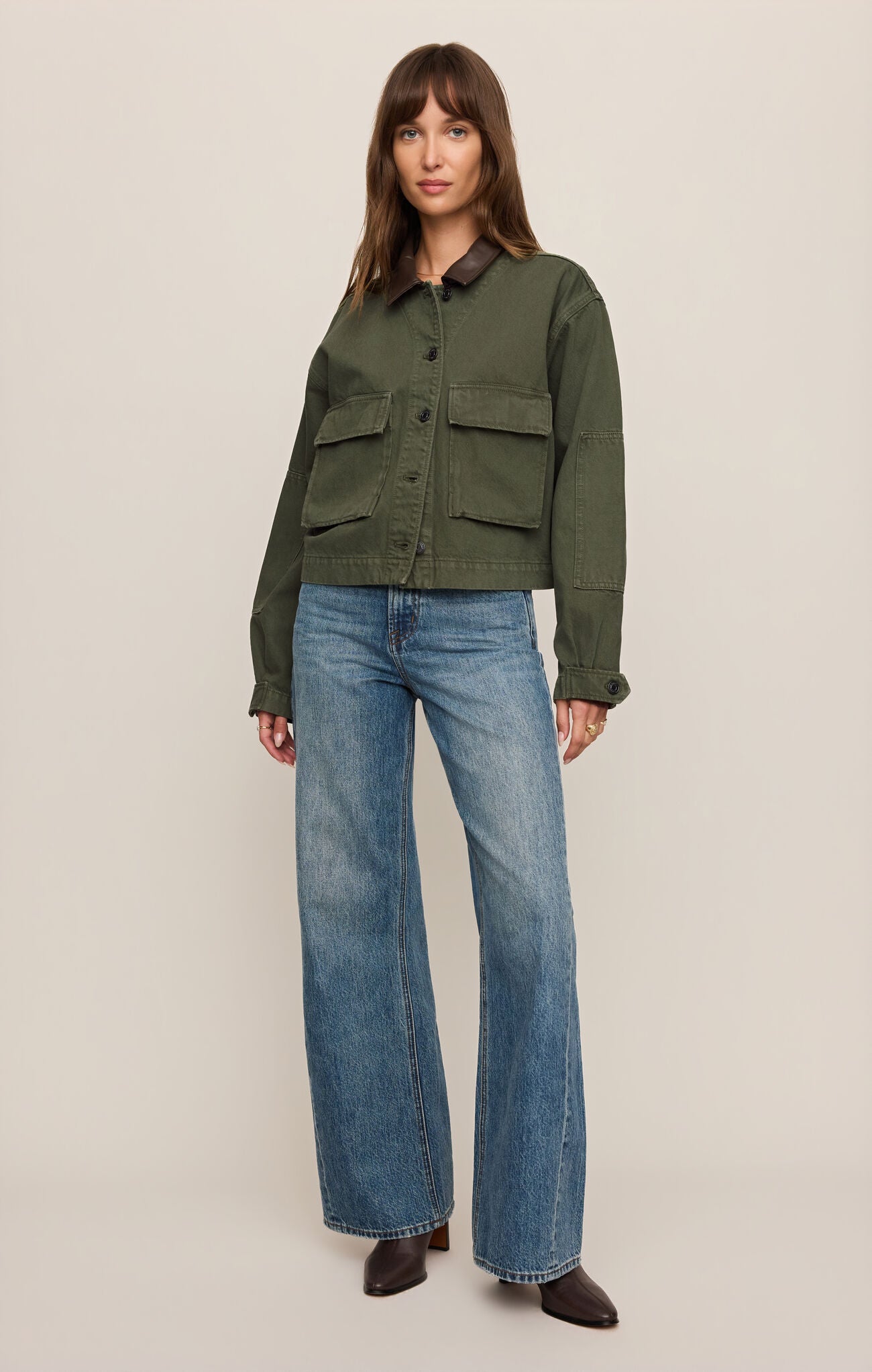 Woman wearing a Dusk Sage Rey Cropped Chore Jacket by z supply with blue jeans and brown boots standing against beige background