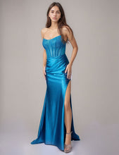 Young woman wearing a teal strapless prom dress with a high slit standing against a plain gray background