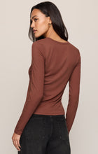 Woman looking over her shoulder in a brown long sleeve shirt and black jeans against a neutral background