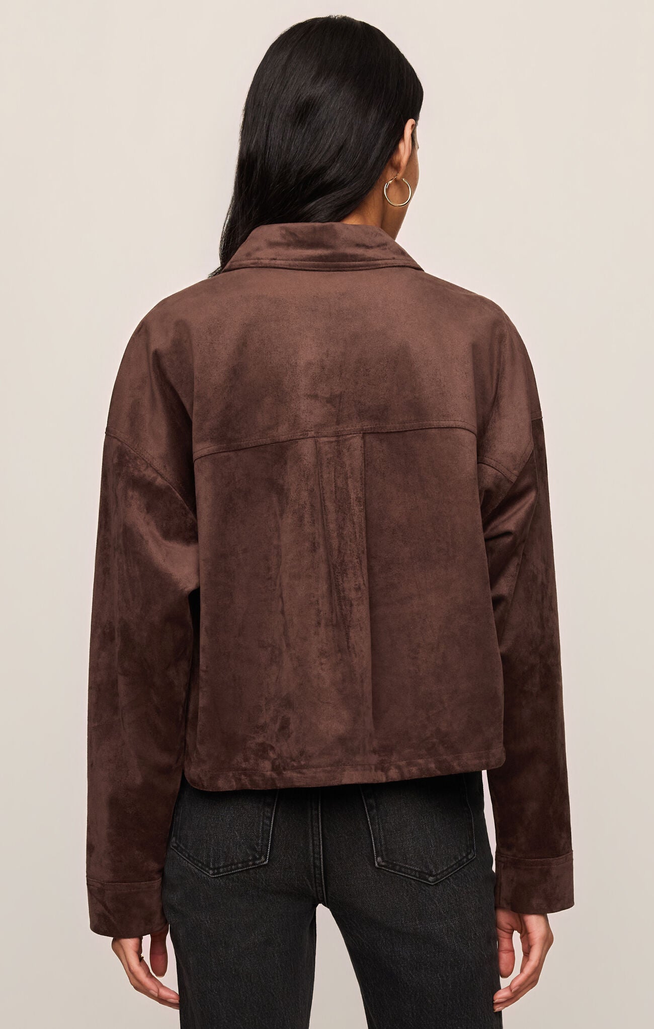 Rear view of a model wearing z supply Dark Chocolate All Day Cropped Suede Jacket with black jeans against neutral background