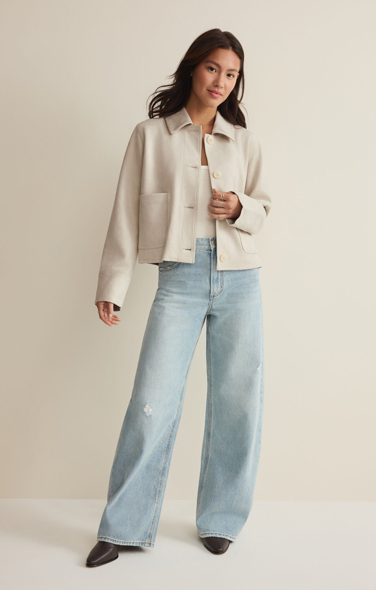Woman with dark hair wearing a cream suede jacket and light blue wide-leg jeans standing against a plain background