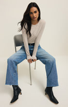 Woman with long dark hair wearing a natural heather fitted long sleeve crew top and wide leg blue jeans sitting on chair