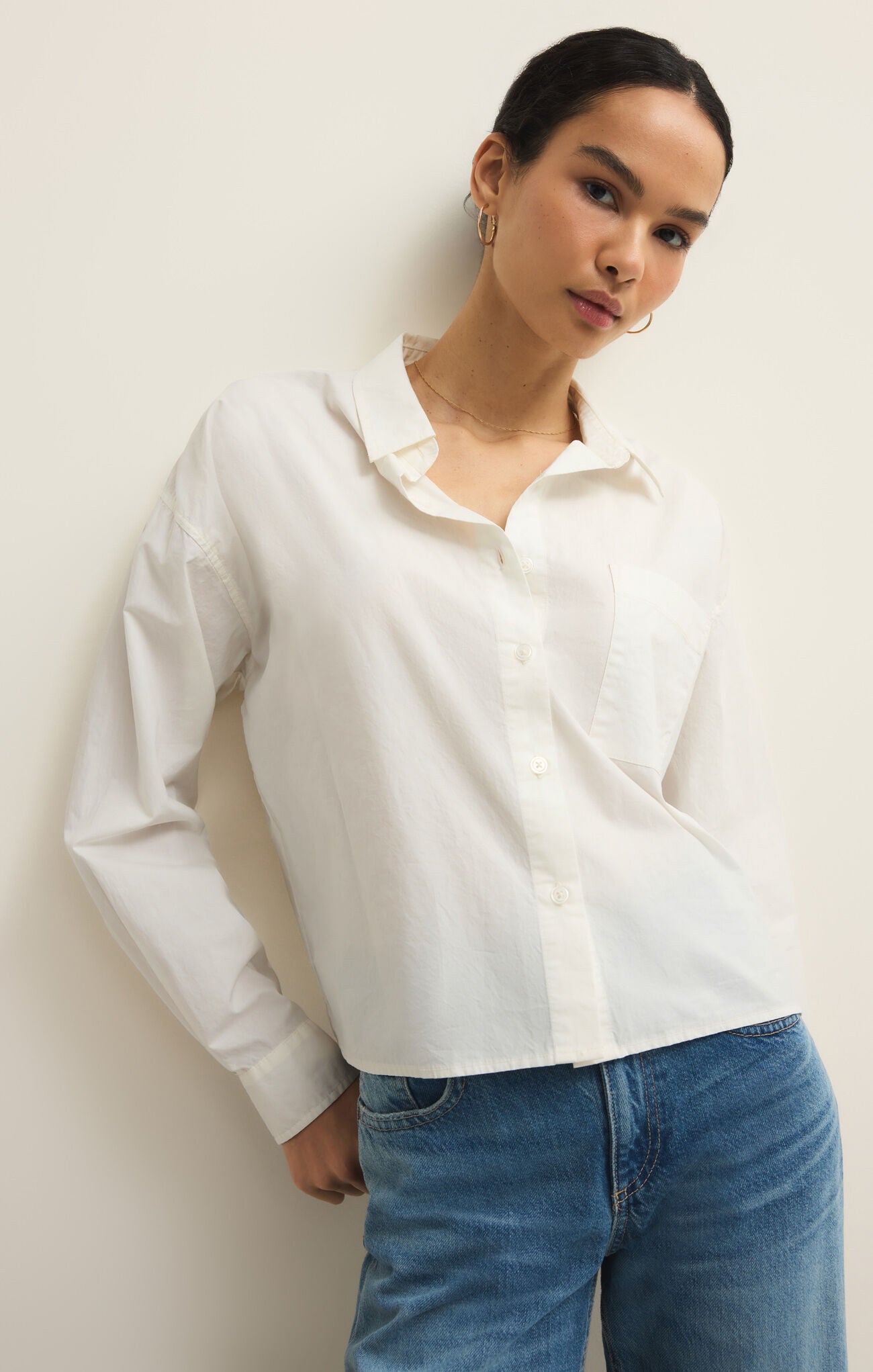 Woman with dark hair wearing a white collared button-up long sleeve shirt and blue jeans standing against a white wall