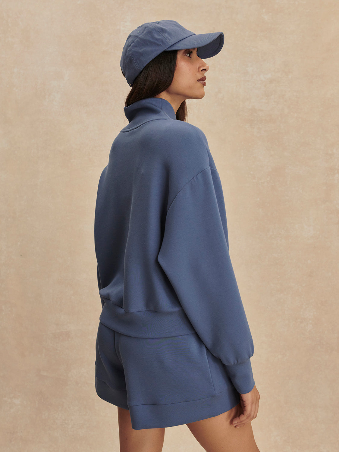 Woman wearing Bijou Blue Davidson Sweat by Varley with matching shorts and cap, posing sideways on beige background