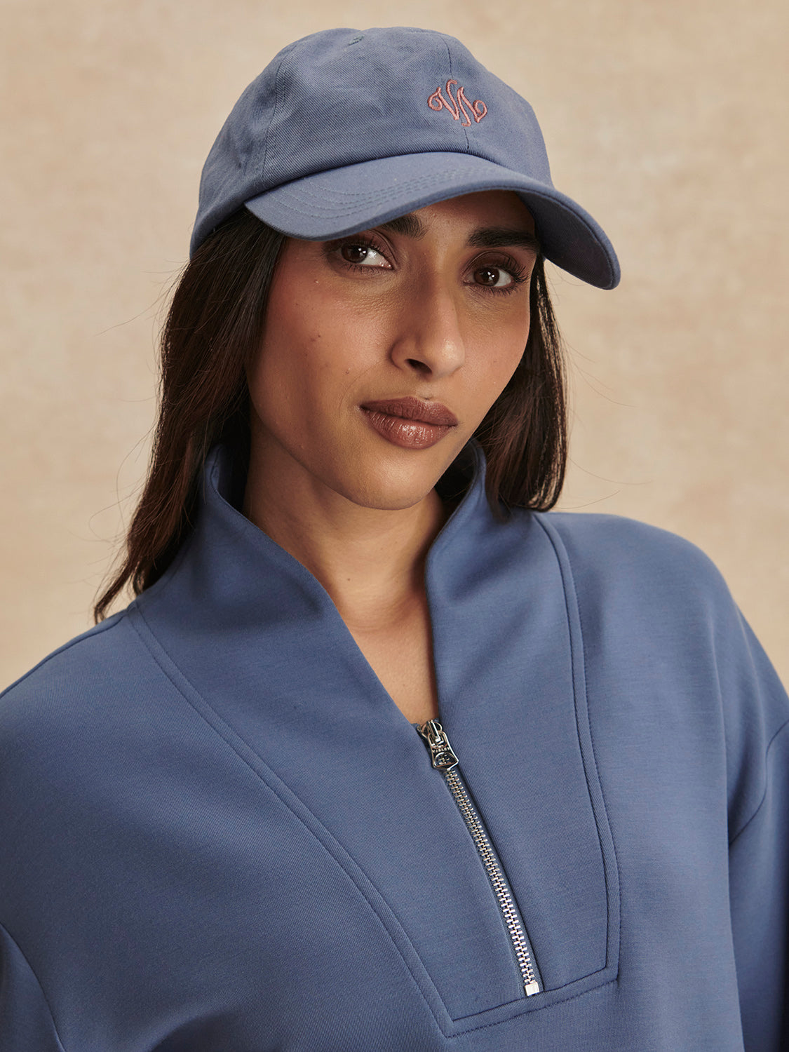 Woman wearing a Varley Bijou Blue Davidson Sweat with a zip collar and matching blue cap against beige background