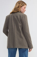 Woman with wavy blonde hair wearing a brown striped blazer and blue pants shown from the back against a plain background.