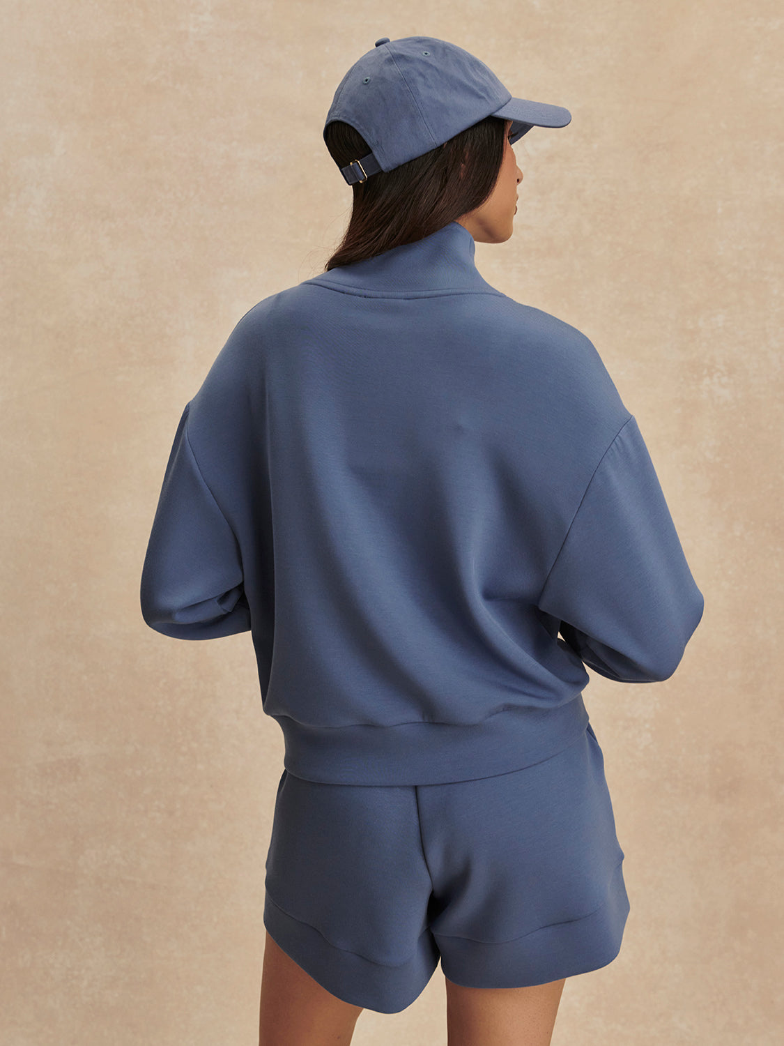 Woman wearing a Bijou Blue Davidson Sweat sweatshirt and shorts set with a matching cap by Varley, shown from the back.