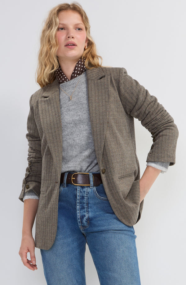 Woman wearing a brown plaid blazer over a gray sweater and blue jeans with a brown belt against a plain background