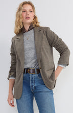 Woman wearing a brown plaid blazer over a gray sweater and blue jeans with a brown belt against a plain background