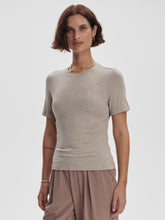 Woman with short brown hair wearing a taupe fitted tee and loose mauve pants standing against a plain background