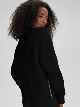 Woman with curly hair wearing a black sweatshirt with a zipper detail on the side standing against a gray background