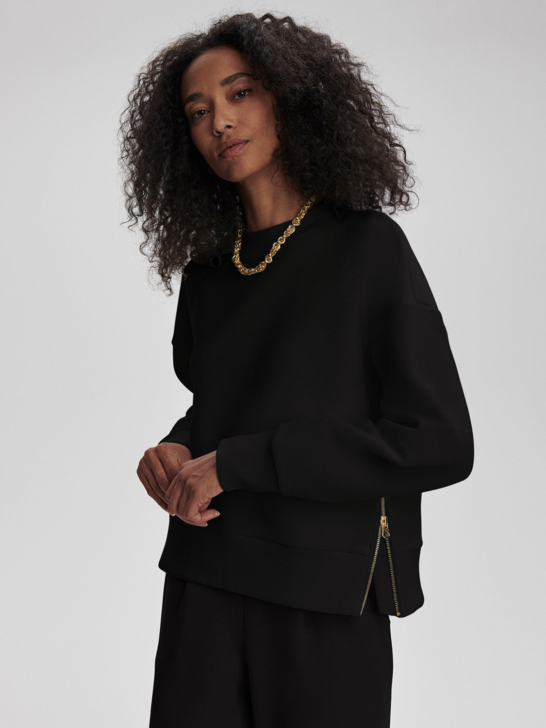 Woman with curly hair wearing a black sweatshirt with side zippers and black pants standing against a gray background